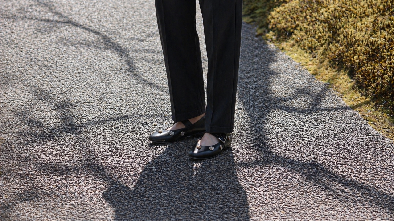 Person wearing Chee’s Charm Mary Jane matte black leather shoes and tailored pants standing on a paved road with Japanese moss